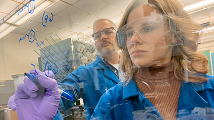 Professor Fair and student wearing gloves and safety goggles draw molecular diagrams on a pane of glass in a lab.