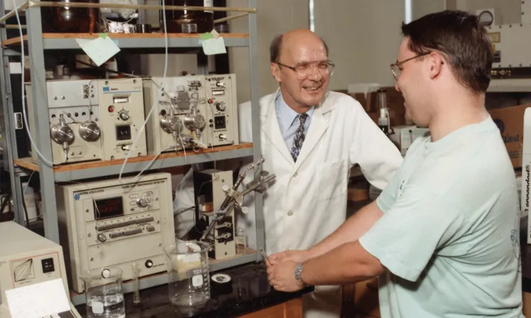a man in a white lab coat talks to a man in a teal shirt in front of a stack of lab equipment