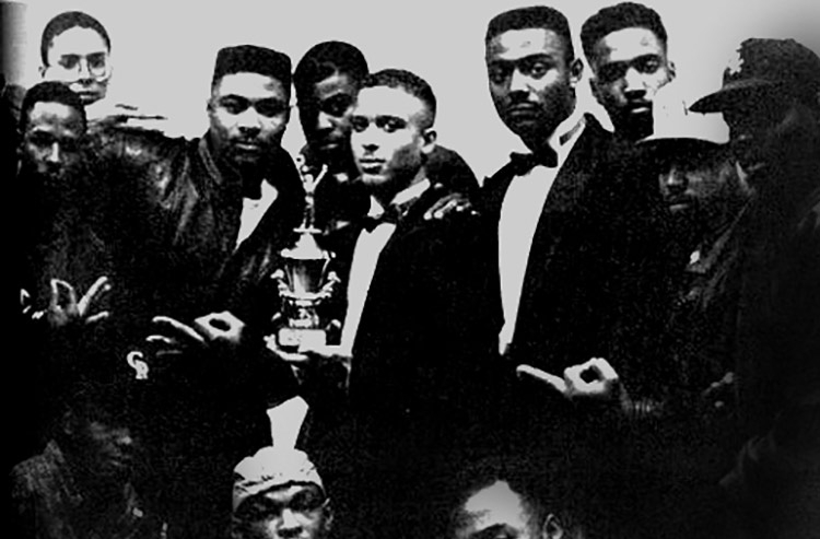 In this black-and-white photo, a group of about 10 casually dressed young men pose around two young men wearing tuxedos, the one in the center holding a trophy. Four men are making a symbolic gesture, forming a circle with their forefinger and thumb and holding the other fingers straight.