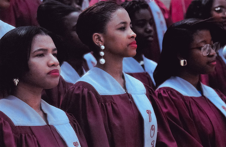 Closeup of three young women within a large group of young women standing at attention in elevated rows, outdoors. All are wearing burgundy, graduation-style gowns with white shoulders and with a white sash with script writing hanging vertically down the left front. 