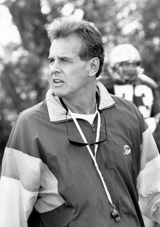 A black-and-white photo of Frank Cignetti in his 50s, wearing an IUP windbreaker with sunglasses and a whistle around his neck, looking to his right from the sideline of a football field