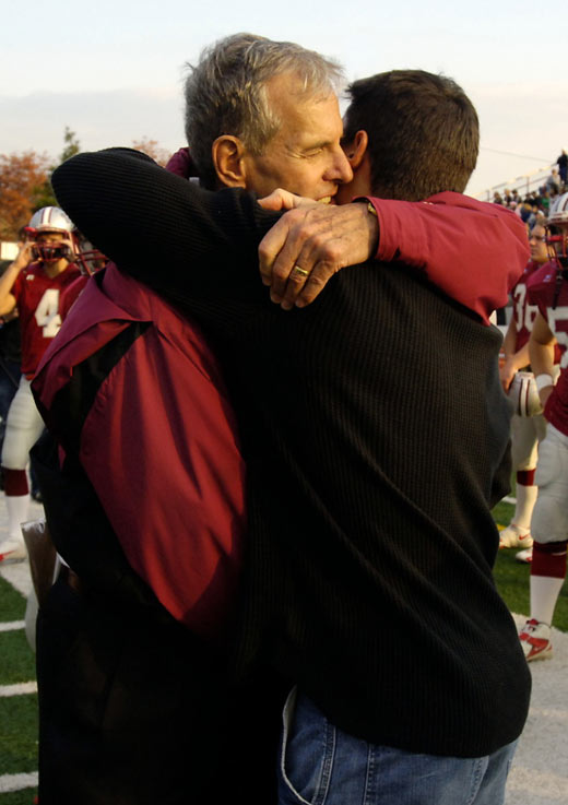 Frank Cignetti, in a dark red sweat suit, hugs a young man with dark hair, a dark shirt, and jeans near the sideline of a football field crowded with players and support staff.