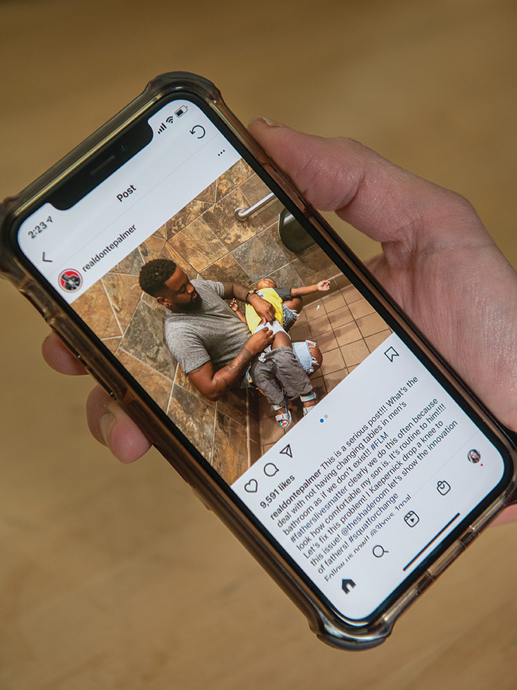 Closeup of a hand holding a mobile phone that displays an Instagram post with a photo of Donte Palmer squatting on the floor of a public restroom with his back against a wall as he changes a toddler’s diaper on his lap