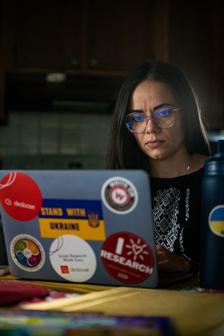A dark-haired woman with glasses works at a laptop computer with six large stickers on the back, including a blue and yellow "Stand with Ukraine" sticker.