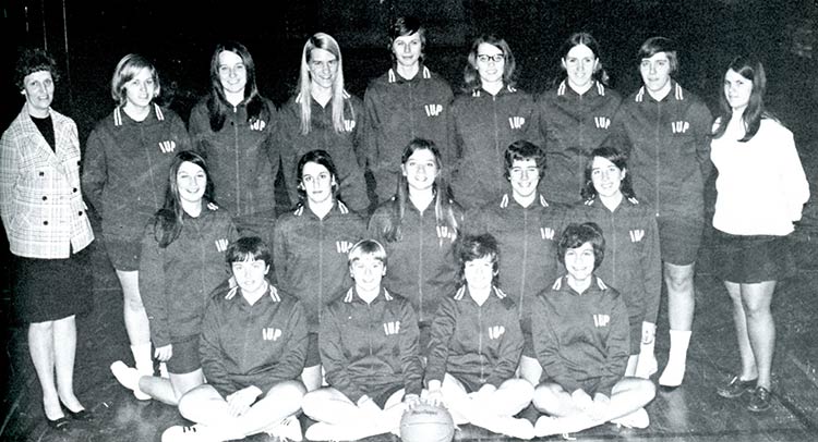 Young women in IUP warm-up jackets pose as a group, with four sitting on the floor in front, five kneeling behind them, and seven standing in back. The two women seated at front center have their hands on a basketball. Two women in dress clothes are standing on either end of the group.