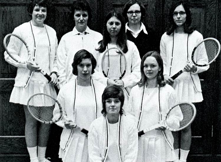 In this black and white photo, eight young women wearing white sweaters, shirts, and tennis skirts hold wooden tennis rackets and pose in an inverted pyramid formation.