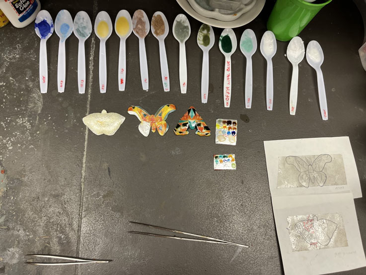 jewelry samples on a table in the process of being painted.  various paint samples sit at the top in plastic spoons