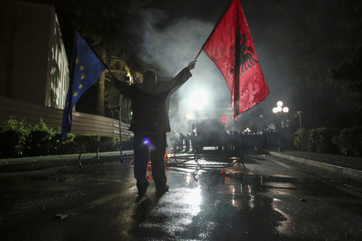 a photo of a man from behind as he holds two flags in the middle of a smoky night time street while facing a police barricade