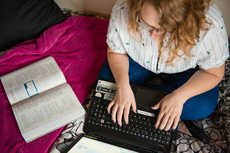 Overhead view of a student sitting on a bed typing on a laptop with an open book laying nearby