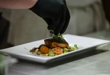 a close up of a finished dish with a hand in a black glove applying a garnish to the top