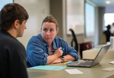 a woman in a blue shirt looking at a student while sitting at a table with a laptop and papers