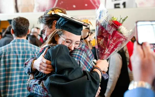 a graduate in cap and gown hugging a family member while holding flowers