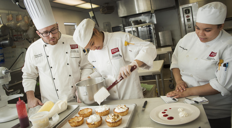Two culinary students watch as a third fills pastry cups with filling.