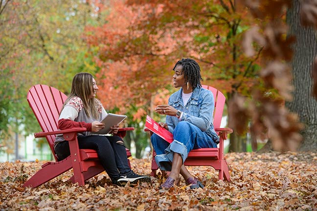 A navigator and a student talk while sitting in Adirondack chairs in the Oak Grove during autumn with fallen leaves on the ground around them.