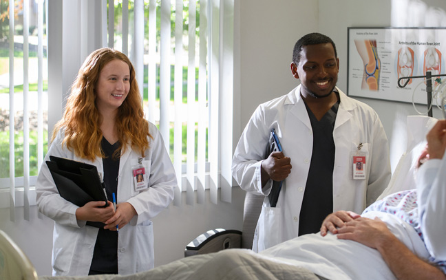 male and female student wearing medical coats stand next to a patient's bed