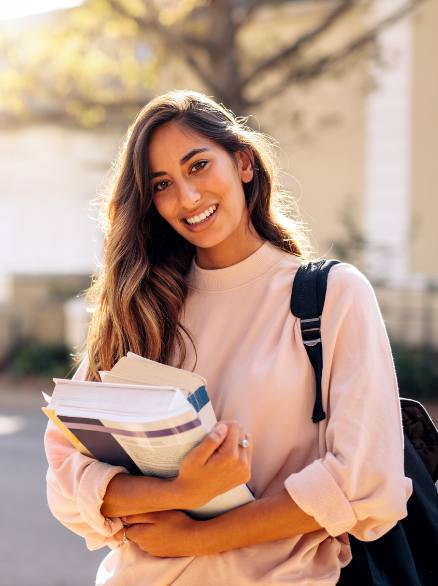 Student with a backpack holding an armload of books and smiling 