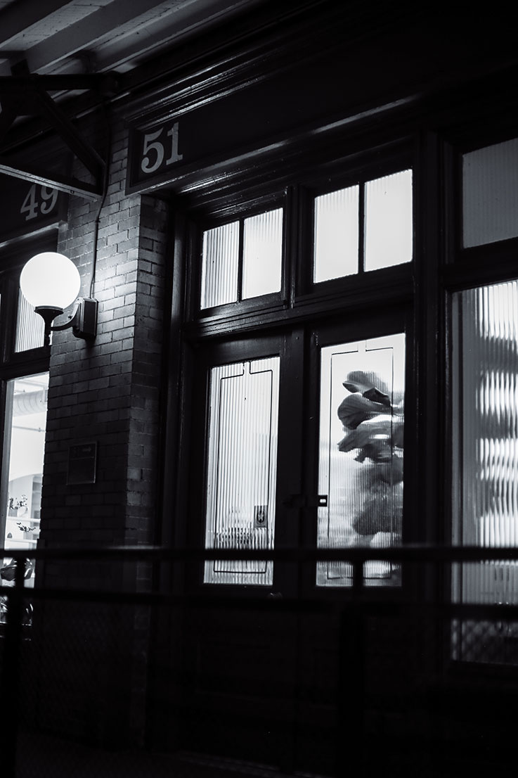 “Grow In Darkness” by Charlotte Smith is a black and white photograph depicting a closed door and lit street lamp. Lights glow from within the building.