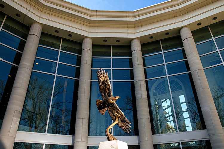 A bronze statue of a hawk in flight, mounted at the top of a white pedestal, is on display in front of a glass-fronted building with columns.