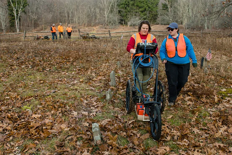 One woman pushes a ground penetrating radar device while another walks alongside her through an old cemetery with sparse, rudimentary gravestones in the fall.