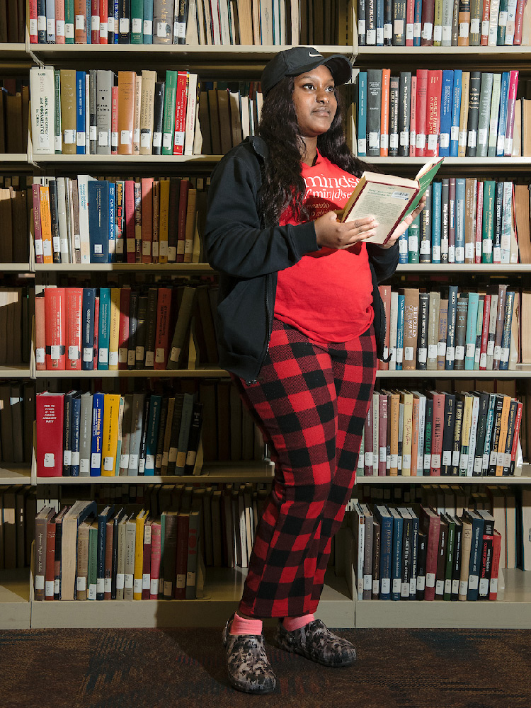 A student wearing red and black plaid pants, a red shirt, and a black jacket reads a book as she stands next to a bookshelf in the library.
