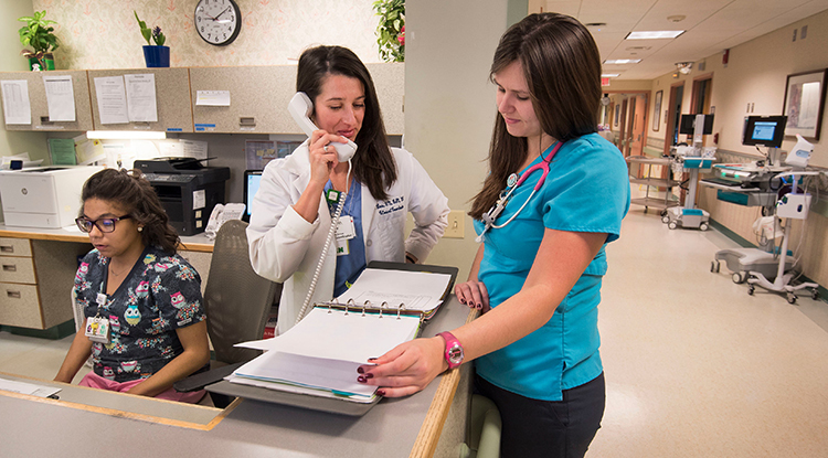 A nurse looking over a binder of information sitting on a counter top while another nurse talks on a phone