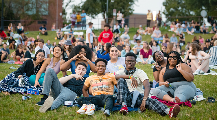 A group of IUP students sitting on the lawn and smiling and posing for the camera, with many other students behind them. 