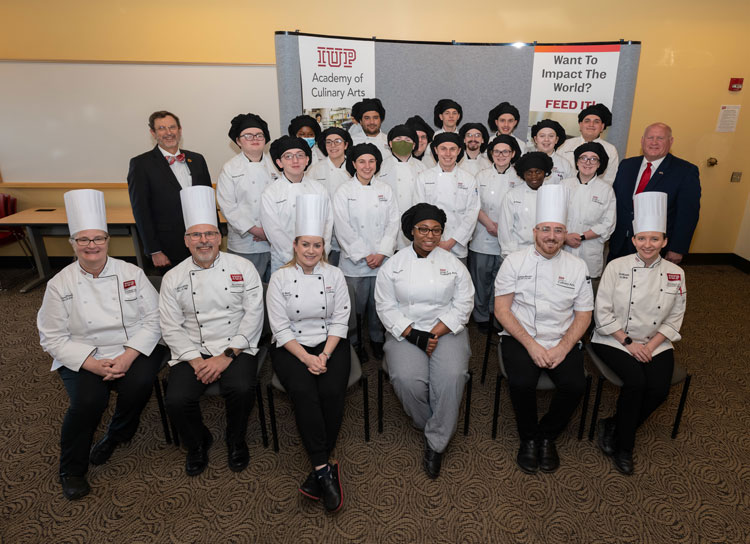 Five culinary faculty members in white chef’s coats and hats and more than 15 culinary students in white chef’s coats and black hats pose in a classroom with IUP President Michael Driscoll on the left and Congressman Glenn Thompson on the right. The faculty members and one student are seated in the front, and the others are standing as a group behind them. 