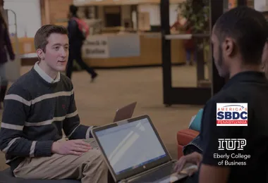 Two people sitting with laptops and facing each other.  SBDC and IUP Eberly Business College logos are on the bottom right.