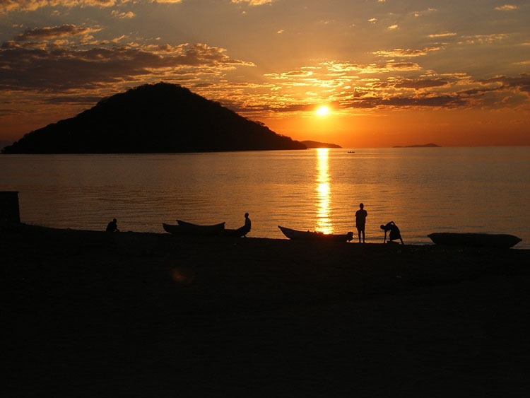 The sun rises or sets over water with people and boats in the foreground The sun rises or sets over water with people and boats in the foreground