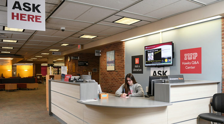 A worker sitting behind a desk with a sign on the wall saying Hawks Q and A Center as well as a banner hanging from the ceiling saying "Ask Here"